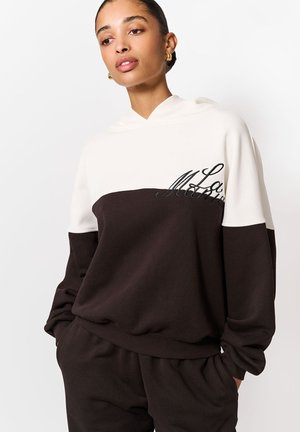 Hanorac cu glugă, cu secțiune superioară crem și secțiune inferioară maro închis. Prezintă brodată inscripția "La" în negru pe piept. Material moale și texturat.