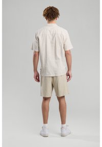 Camicetta a maniche corte a righe beige con yoke posteriore, abbinata a pantaloni beige. Il modello indossa sneakers bianche, in piedi su uno sfondo chiaro.