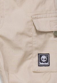 Tissu beige avec une finition texturée, comportant un accent vert sur le rabat de la poche. Affiche un patch du logo Timberland noir sur la poche.