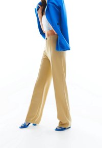 Pantalons beige à jambes larges avec une taille haute, associés à un top blanc court et un blazer bleu vif. Sandales bleues à semelle plate.