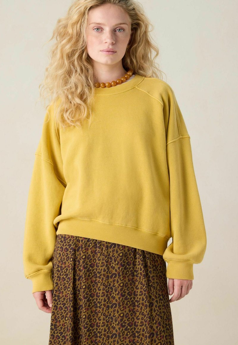 Personne jeune avec de longs cheveux blonds bouclés portant un sweatshirt oversize jaune moutarde, une jupe imprimée léopard marron, et un collier de perles ambre.