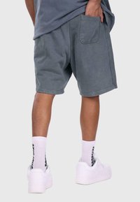 Grå bomuldsshorts med en baglomme, iført hvide sokker med sort tekst og hvide sneakers, set fra baghovedet.