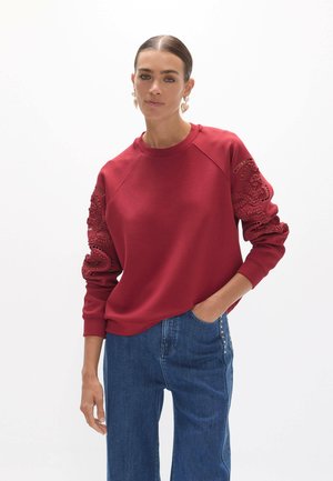 Mujer con el pelo peinado hacia atrás lleva sudadera roja con detalles de encaje en las mangas y vaqueros azules con la mano en el bolsillo frente a un fondo blanco.