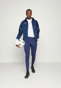 Veste et pantalon de sport bleu marine avec des bandes latérales blanches, associés à une chemise blanche et des chaussures de foot noires, tenant un ballon de soccer.