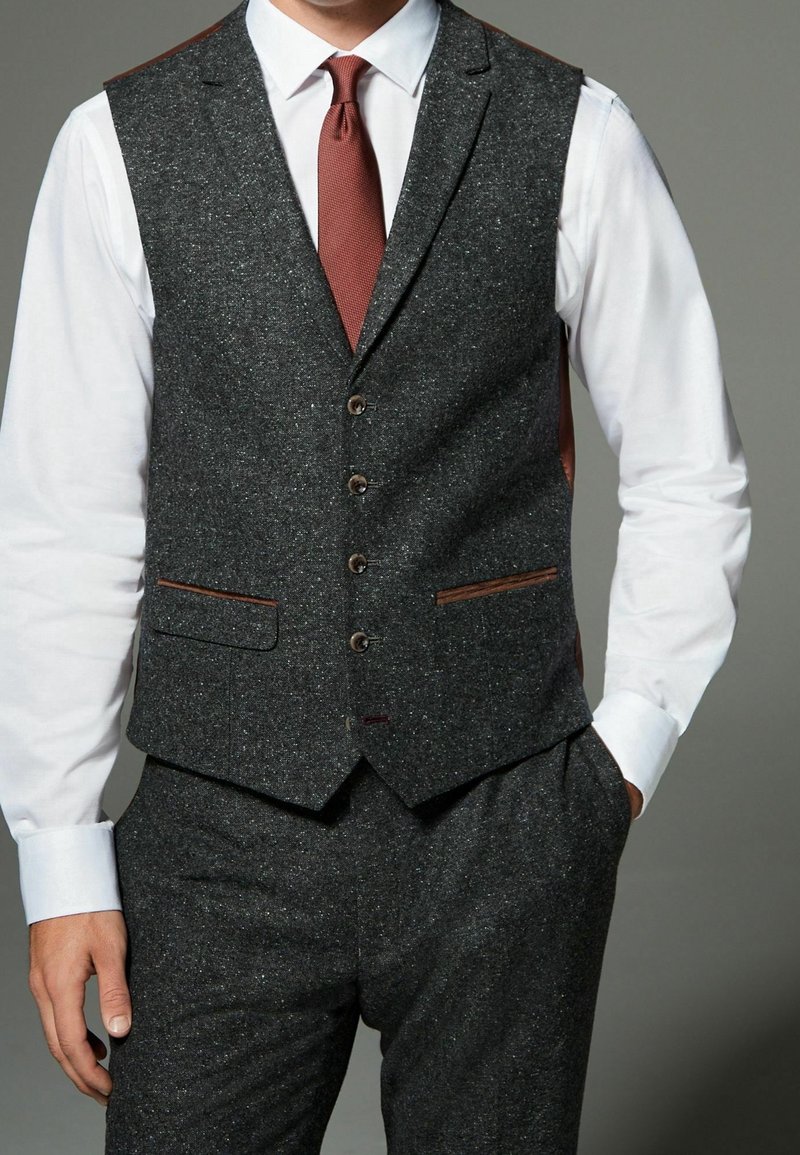 Homme portant un gilet en tweed gris foncé et un pantalon assorti, une chemise blanche et une cravate marron texturée, une main dans la poche.