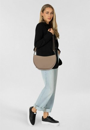 Femme aux longs cheveux blonds portant un blazer noir, un jean bleu clair retroussé, des baskets compensées noires, et un sac à bandoulière beige.