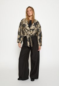 Beige a čierna kvetinová vzorovaná bunda s dlhým rukávom, previazaná v páse, v kombinácii s čiernymi nohavicami s vysokým pásom a širokými nohavicami a čiernymi topánkami.