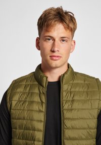 Gilet invernale verde oliva con colletto alto, texture trapuntata e zip frontale. Indossato sopra una maglietta nera. Materiale morbido e leggero.