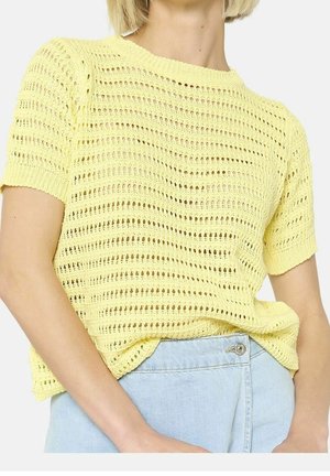 Kvinde iført en kortærmet, lysegul striktrøje med åbent mønster, stukket ned i højtaljede lyseblå denimjeans.