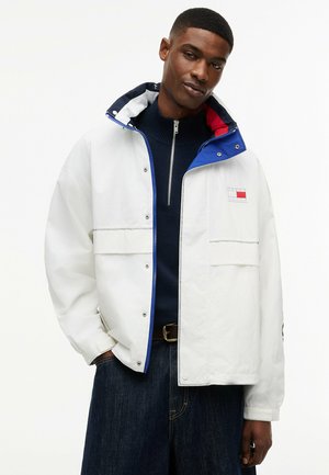 Mand iført hvid hættetrøje med blå og rød indvendig foring over en marineblå zip-up sweater og mørke jeans, stående mod en hvid baggrund.