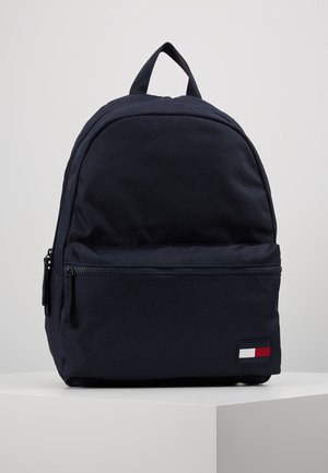 Granatowy plecak z przednią kieszenią na zamek oraz prostokątną naszywką z logo w kolorach czerwonym, białym i granatowym w dolnym prawym rogu.