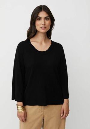 Top nero a maglia con scollatura rotonda ampia, maniche a tre quarti e vestibilità comoda. Indossato con pantaloni beige e un bracciale rigido dorato.
