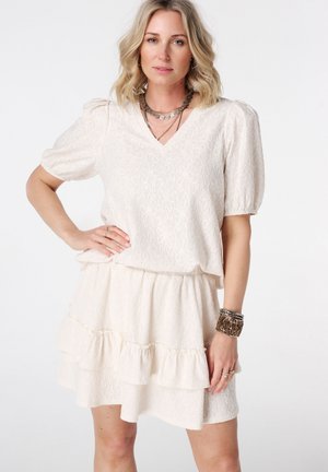 Femme aux cheveux blonds portant une robe blanche texturée avec des manches bouffantes, une jupe à volants et plusieurs bracelets au poignet droit sur fond blanc.