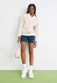 Μπέζ pullover μπουφάν με λευκό γιακά, ναυτικές βερμούδες ποδηλάτου, λευκά αθλητικά παπούτσια και κρεμ τσάντα; λεία υφή και μίνιμαλ σχέδιο.