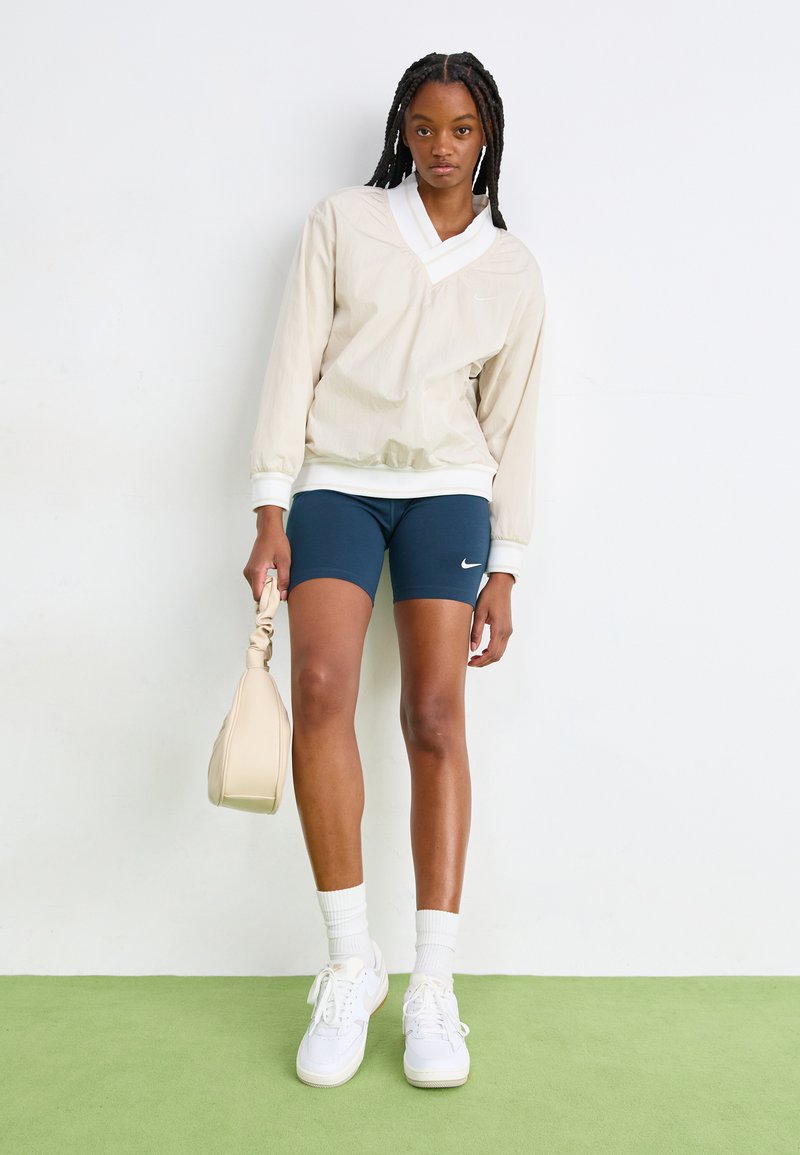 Beżowa kurtka w pulowerze z białym kołnierzykiem, granatowe szorty kolarskie, białe sneakersy i kremowa torebka; gładka tekstura i minimalistyczny design.