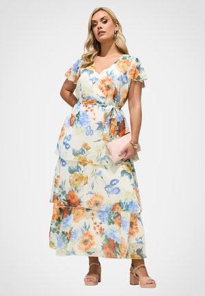 Kvinde iført en blomsterprintet, lagdelt midi-kjole med flagrende ærmer, der holder en støvet rosa clutch og bærer nude blokhælede sandaler.