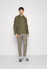 Jachetă din bumbac verde măsliniu cu multiple buzunare frontale, purtată peste un tricou asortat. Pantaloni gri cu carouri și adidași cu accente albastre.