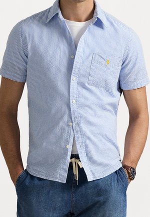 Homme portant une chemise à manches courtes à rayures bleu clair et blanches par-dessus un t-shirt blanc, avec un pantalon bleu et une montre-bracelet, les mains dans les poches.