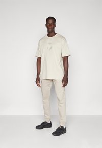 Beige Baumwoll-T-Shirt mit einem dezenten Logo, kombiniert mit beigen Jogginghosen. Schwarze Schuhe mit einer strukturierten Sohle runden das Outfit ab.
