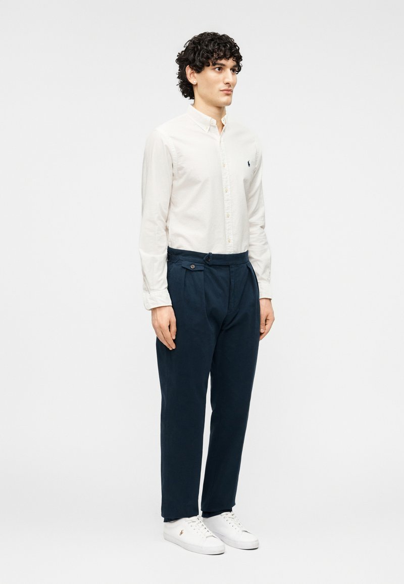 Chemise claire à boutons avec col ; pantalon bleu marine plissé ; baskets blanches avec une marque discrète ; tissu lisse et coupe décontractée.