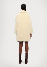 Cappotto in finta pelliccia shearling color crema, morbido e fluffy, con un ampio colletto, vestibilità oversize e lunghezza fino a metà coscia, abbinato a stivali marroni al ginocchio.