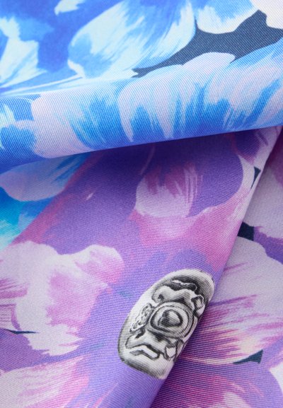 Gros plan sur un tissu plié avec des motifs floraux bleus et violets et un petit motif de masque de style aztèque en noir et blanc.