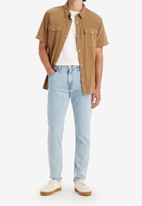 Man som bär ljusblå jeans, vita sneakers, vit t-shirt och öppen brun kortärmad skjorta med knappar, står med ena handen i fickan.