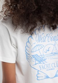 Valkoinen puuvillapaita sinisellä graafisella designilla, jossa on ranta-alue, tuoli, aurinkovarjo ja teksti: "Länsirannikon Yacht Club Kalifornia."