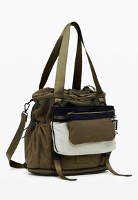Sac à épaule vert olive avec un cordon de serrage en haut, des accents en maille, des poches zippées à l'avant en noir et crème, et des bretelles réglables pour plus de polyvalence.