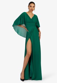 Smaragdgrünes Kleid mit schwingenden Ärmeln, gerafftem Oberteil, tiefem V-Ausschnitt und hohem Schlitz. Glatter Stoff, bodenlanger Schnitt, kombiniert mit schwarzen Absätzen.