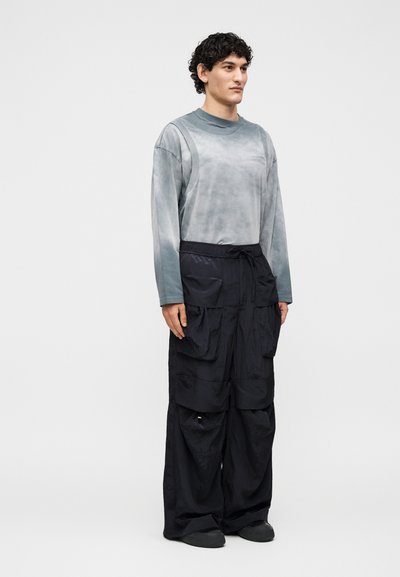 Νέος άντρας που στέκεται, φοράει γκρι μπλούζα μακρυμάνικη και φαρδιά μαύρη παντελόνια cargo με πολλές τσέπες σε απλό λευκό φόντο.