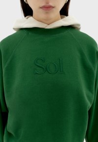 Zelená mikina s bílou kapucí, na přední straně má vyšité logo "Sol". Měkká textura, žebrovaný límec a manžety, raglánové rukávy.