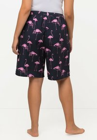 Una persona sta in piedi a piedi nudi, indossando pantaloni corti blu navy con stampa di fenicotteri rosa e una maglietta bianca, vista di spalle su uno sfondo neutro.