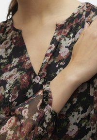 Blusa nera floreale con scollatura a V profonda. Presenta maniche lunghe trasparenti con polsini arricciati. Il motivo floreale include fiori rosa e bianchi su uno sfondo scuro.