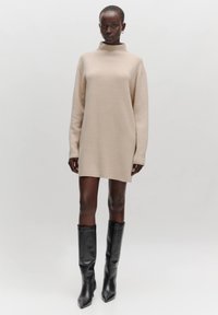Beige oversized trui-jurk met een hoge hals, lange mouwen en een geribbelde textuur, gecombineerd met zwarte knie-hoge stiletto-laarzen.