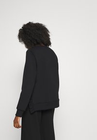 Personne avec des cheveux noirs bouclés portant un haut à manches longues noir et un pantalon noir, tournée de dos devant un fond blanc uni.