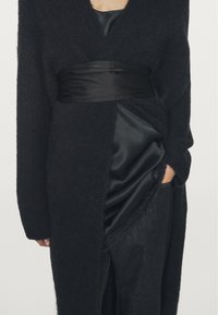 Czarny puszysty, oversized kardigan z głębokim dekoltem w serek, zawiązany w talii błyszczącą czarną satynową wstążką, nałożony na satynową bluzkę i ciemne spodnie.