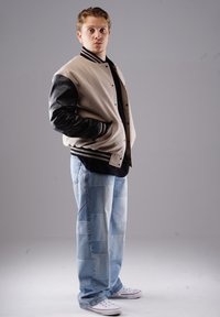Beige- und schwarze Collegejacke mit Druckknöpfen, kombiniert mit einer mit Patches verzierten hellblauen Jeans und weißen Sneakers. Das Modell steht im Profil.