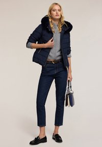 Námořnická prošívaná bunda s kožešinovou kapucí, šedý pletený svetr, tmavé džíny a černé mokasíny. Drží vícebarevnou kabelku.