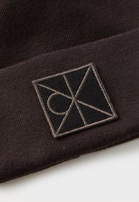 Beanie lavorato a maglia marrone con una toppa quadrata nera e logo geometrico ricamato. Texture morbida e dettaglio a coste.