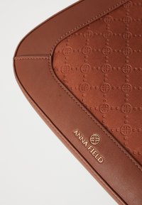 Bolsito de cuero marrón con patrones circulares en relieve y logo dorado "ANNA FIELD" en el borde de cuero liso.