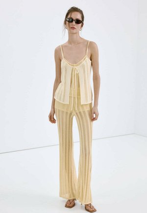 Woman wearing pale yellow knit sleeveless top and matching wide-leg pants, dark sunglasses, standing against plain white background.