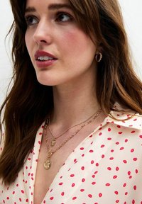 Goudkleurige gelaagde kettingen met muntbedels, gedragen over een lichte blouse met een rode lippenafdruk. Gladde textuur en een delicate kettingontwerp.