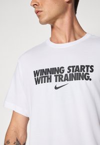 Hvit T-skjorte med svart tekst som sier "VINNING STARTER MED TRENING" og Nike-logoen. Korte ermer, rund hals, laget av bomull.