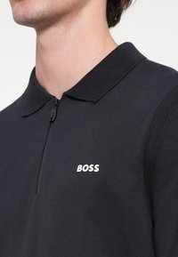 Nāvijas polo krekls ar rāvējslēdzēju apkaklē, raksturīga teksturēta auduma un baltu "BOSS" logo kreisajā krūtī.