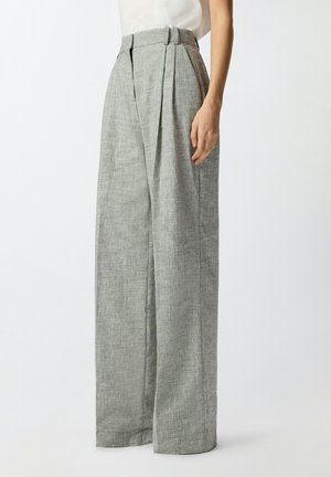 Mujer con pantalones grises de tiro alto y pierna ancha con pliegues frontales, combinados con una blusa blanca sin mangas, parada frente a un fondo liso.