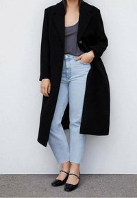 Cappotto lungo nero con un design sartoriale, maglia grigia, jeans a vita alta azzurri chiari e ballerine nere con cinturini alla caviglia; texture minime.
