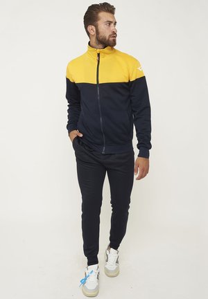 Mann mit Bart in einer marineblauen und gelben Jacke mit Reißverschluss und schwarzer Hose, steht mit einer Hand in der Tasche und schaut zur Seite.