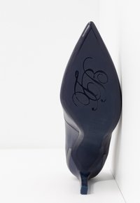 Chaussure à talon haut bleu marine avec un bout pointu, présentant une surface texturée et un petit talon. La semelle comporte un logo et des détails décoratifs embossés.