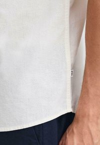 Camisa de lino blanco ligera con un acabado texturizado sutil. Presenta una etiqueta en la costura lateral y un dobladillo ligeramente curvado para un ajuste relajado.